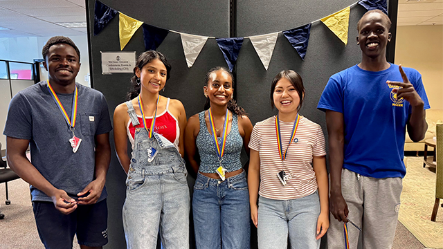 Five students with colorful lanyards stand together in an office setting.