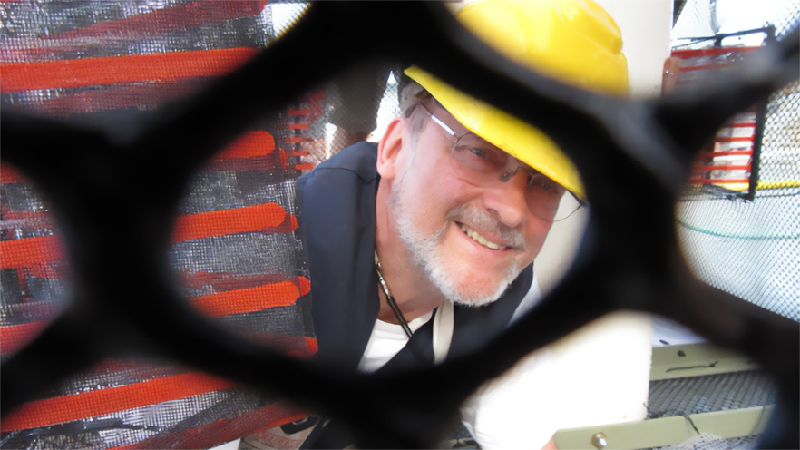Paul Yancey, wearing a yellow hard hat, is seen smiling through a hole in black netting in the foreground.