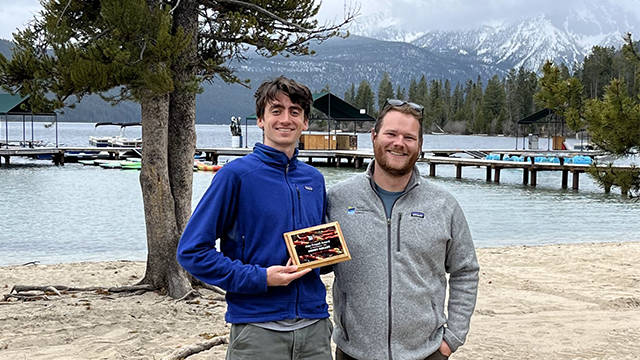 Henry Roller ’25 (left, holding an award) and Mitch Cutter ’17 (right) stand on a sandy beach with a body of water and snowcapped mountains in the background.