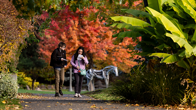 Two students walk across campus with fall colors visible on the trees in the background.