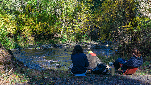 Two students sit next to a stream with nearby books and backpacks.
