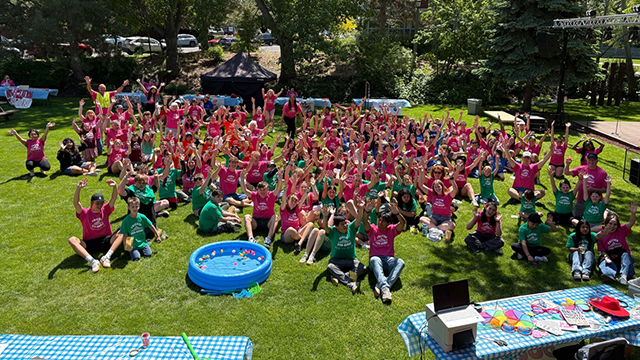 A large crowd of people wearing matching pink or green shirts, seated in a grassy area, waving their hands in the air.