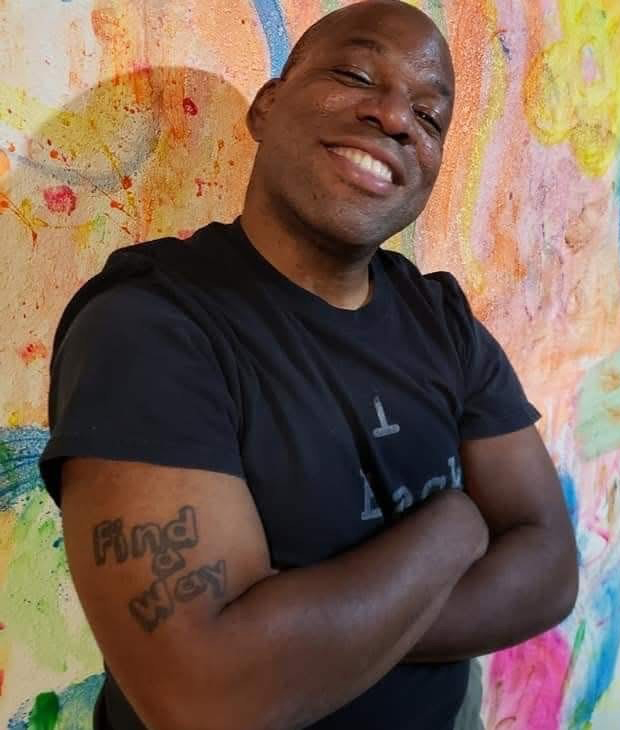 Portrait of Nathan Brannon, arms folded, standing in front of a wall with colorful abstract painted designs.