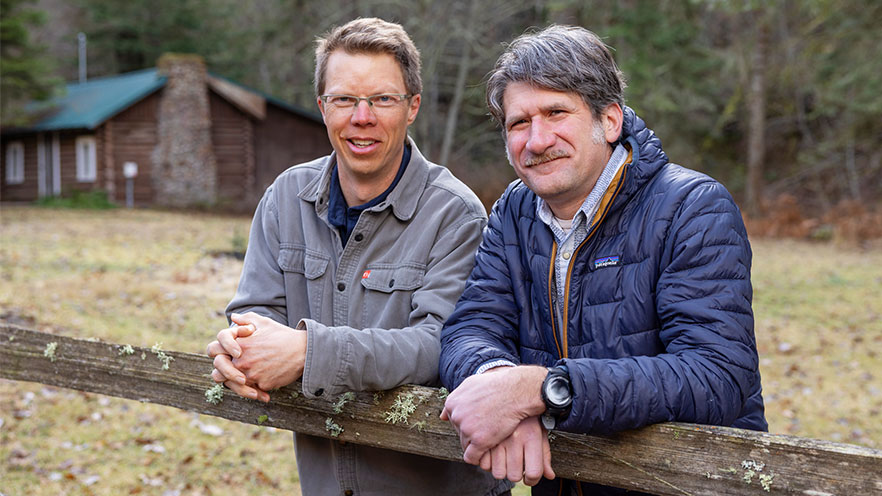 Two professors lean against a wooden fence with a cabin and trees visible in the background.