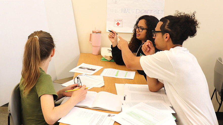 Three students work together at a table strewn with papers.