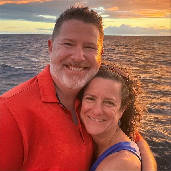 Two people embracing each other and smiling at the camera. There is a body of water behind them and a sunset happening in the distance 