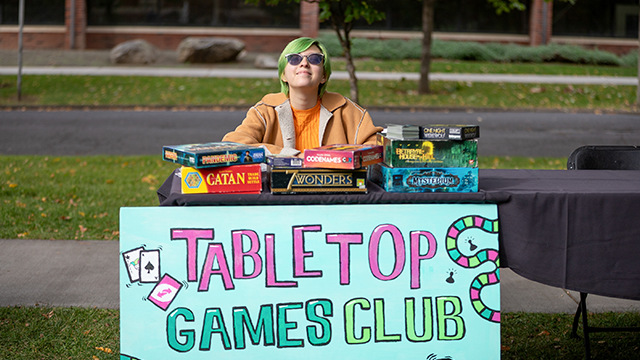 A person with green hair and wearing sunglasses sits behind a table with stacks of board games and a sign that reads “Tabletop Games Club”