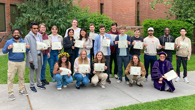 A large group of students and faculty stand outside Olin Hall, each person holding a certificate.