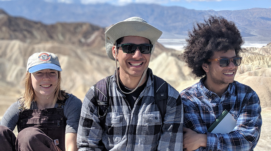 Three students pose together outdoors, wearing hats and sunglasses.