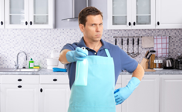 Serious man wearing a blue apron and gloves holding a spray bottle in kitchen.