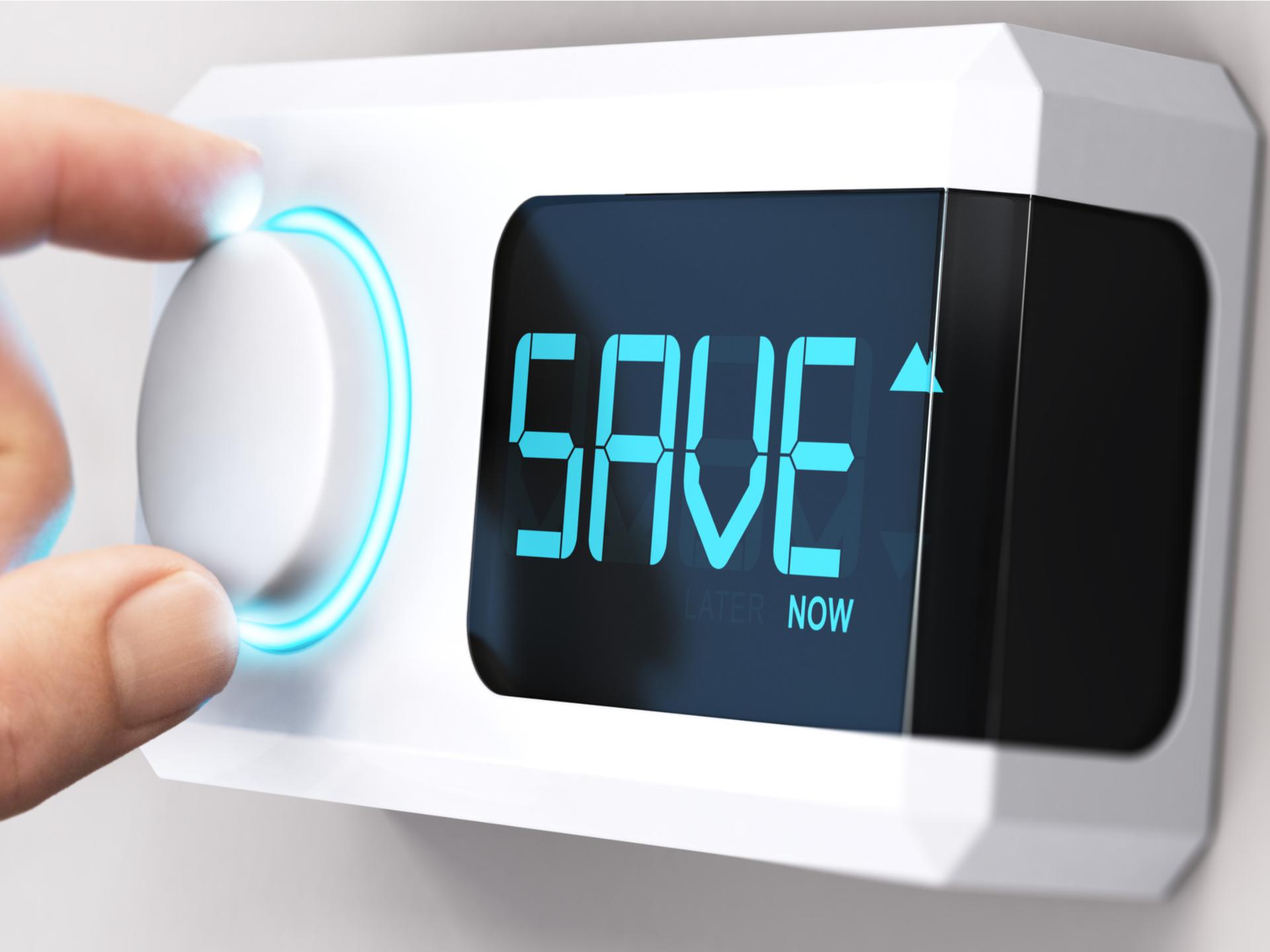 Close up photo of a hand turning down a dial on a thermostat with the word, “SAVE” on the thermostat digital screen