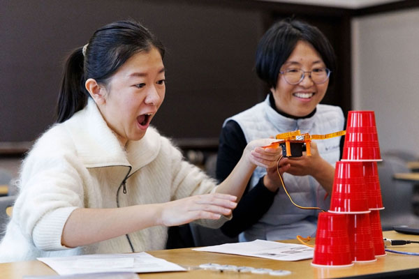 Two women use an orange bionic claw to build a pyramid of red Solo cups