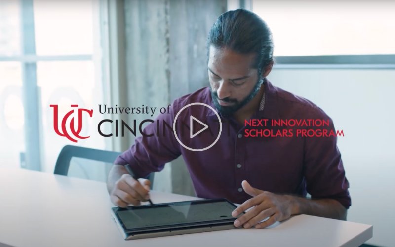 Man writing on a tablet at a desk with “University of Cincinnati” and “Next Innovation Scholars Program” text overlayed, with a play button in the center.
