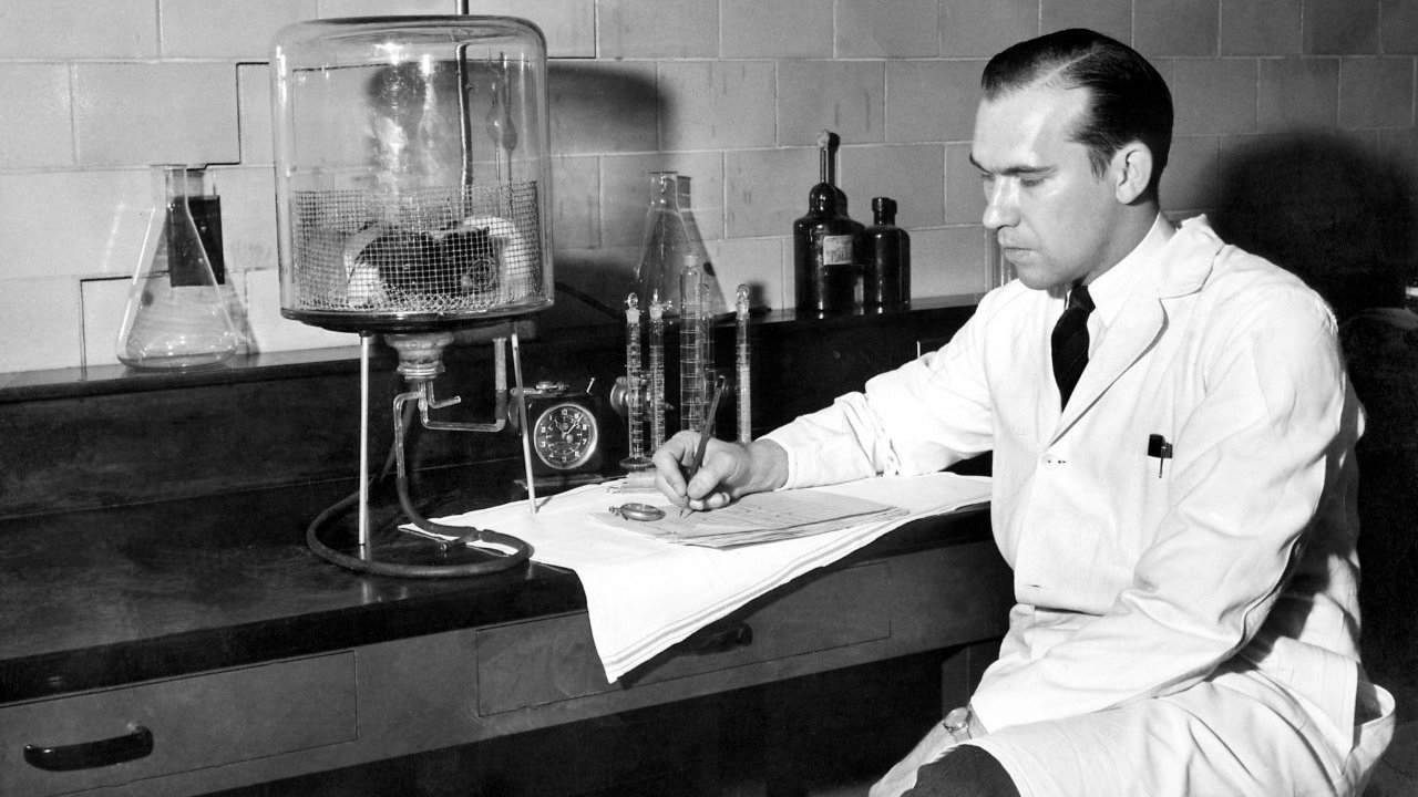 George Rieveschl in his lab coat sitting at a desk, writing notes beside glass lab equipment and flasks