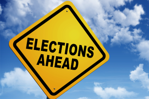 A yellow road sign with black text that says Elections Ahead.