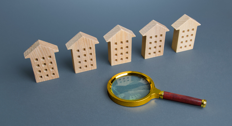“Magnifying glass examining a row of small wooden house models, representing closer scrutiny of homes in today’s housing market.” “Magnifying glass examining a row of small wooden house models, representing closer scrutiny of homes in today’s housing market.”