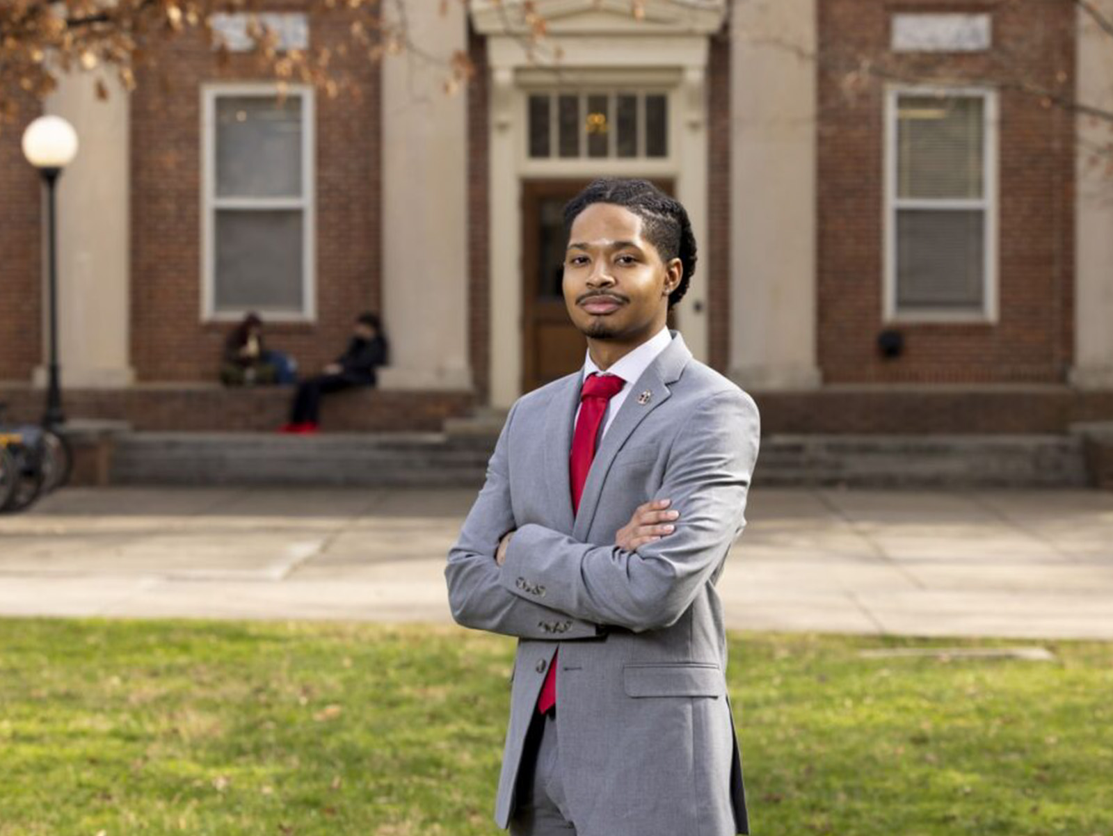 Corey Staughter poses with his arms crossed in a gray suit in front of a historic building on UGA's north campus. 