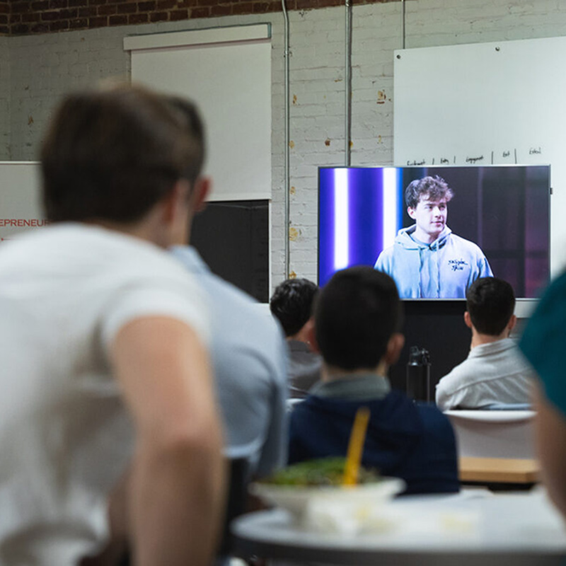 A crowd watches Skunk Skins founder Matt Tesvich on a TV during a debut party for the Amazon Prime show Buy It Now.