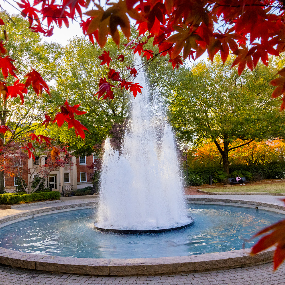 View of the Herty Field fountain with sunlight filtering through trees full of colorful fall leaves.