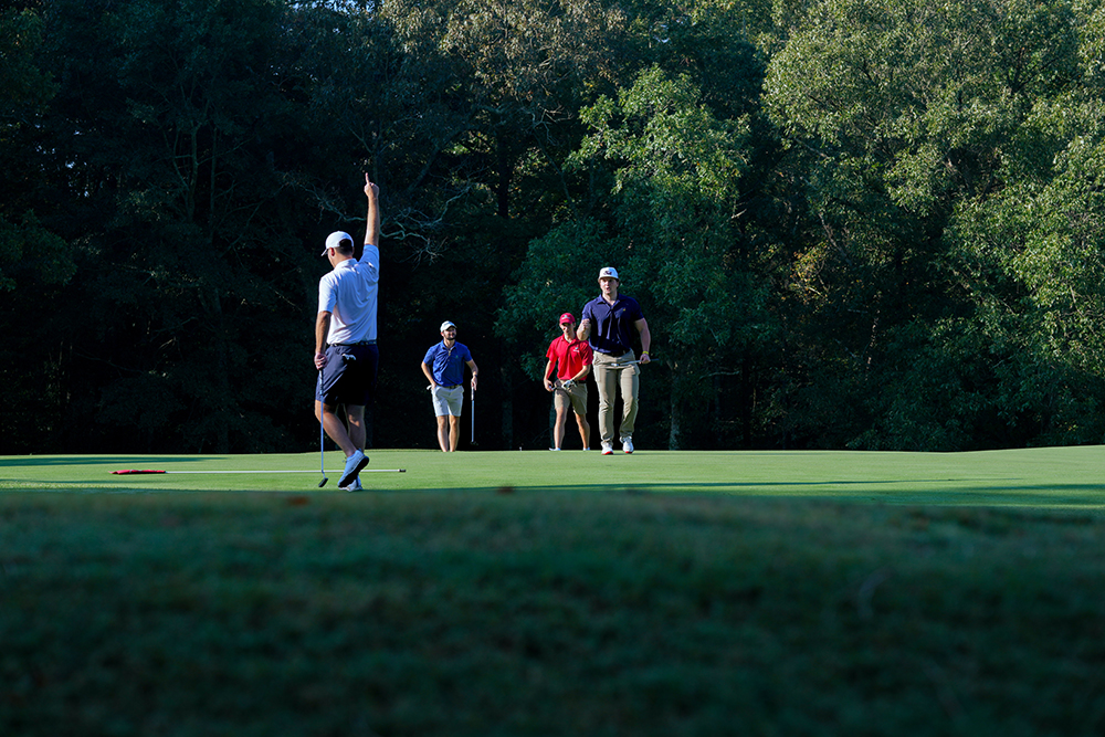 A group of 4 golfers. The closest golfer from the left holds his right arm and points to the sky mid stride