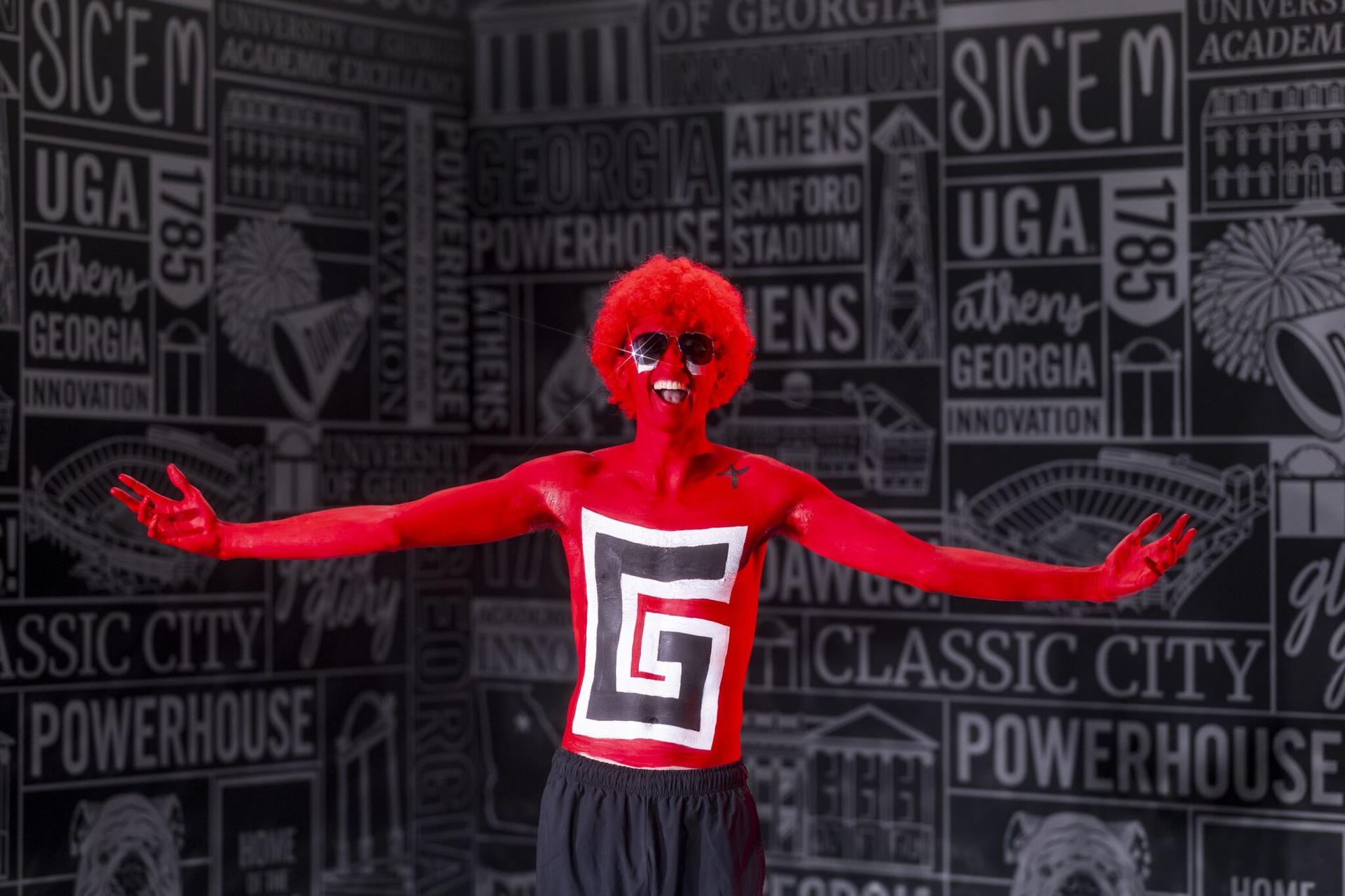 JT Forsyth, wearing sunglasses and a red wig, covered fully in red body paint, with a huge painted G covering his torso, poses for a photo