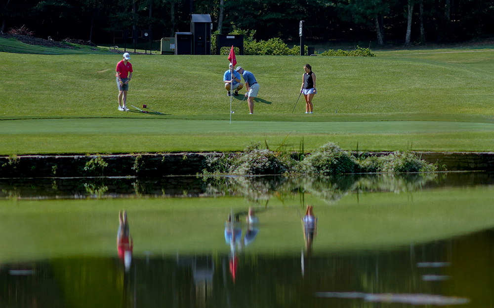 Long shot from across a body of water of 4 golfers playing just outside the putting green across.