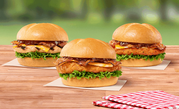  Smokehouse BBQ Bacon Sandwich with Grilled filet, Smokehouse BBQ Bacon Sandwich with Original filet, Smokehouse BBQ Bacon Sandwich with Spicy filet sitting on a table next to a red and white napkin with grass and trees in the background