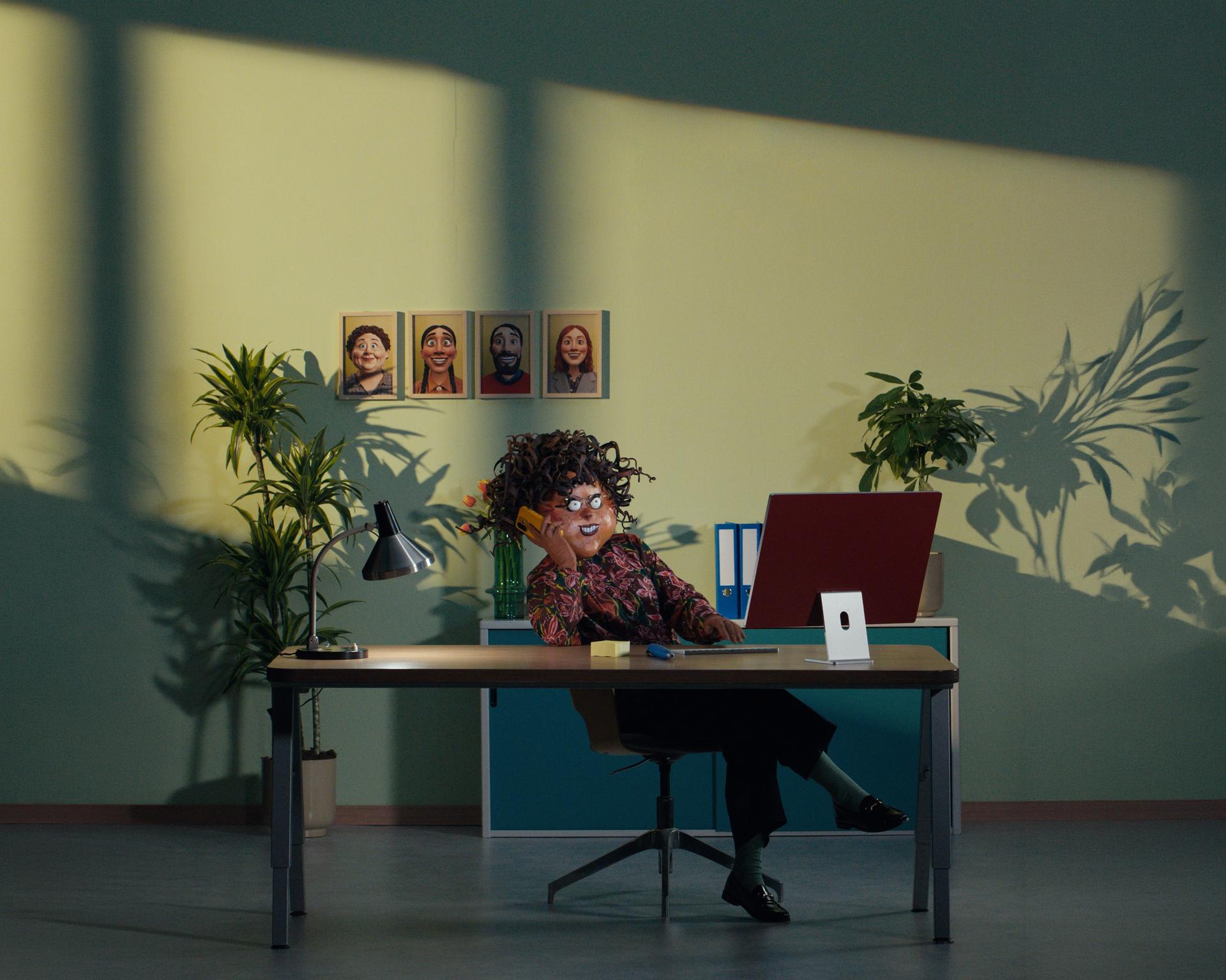 Beeld uit campagnevideo: vrouw zit achter een bureau in een kantoor met planten, een laptop en portretten aan de muur.