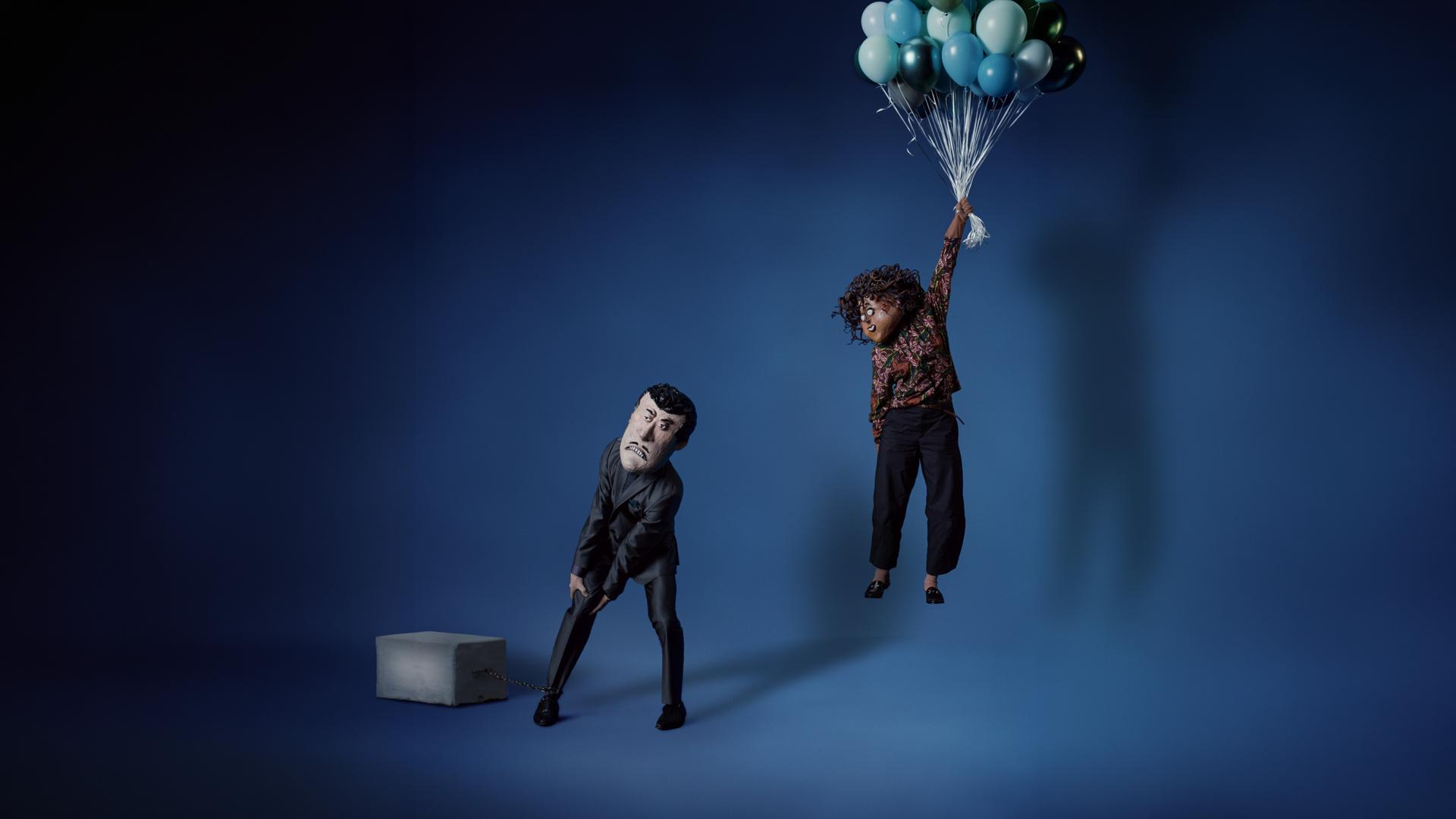 Beeld uit de video 'Balonnen': Twee personen staan in een donkere ruimte; één houdt een tros blauwe ballonnen vast.