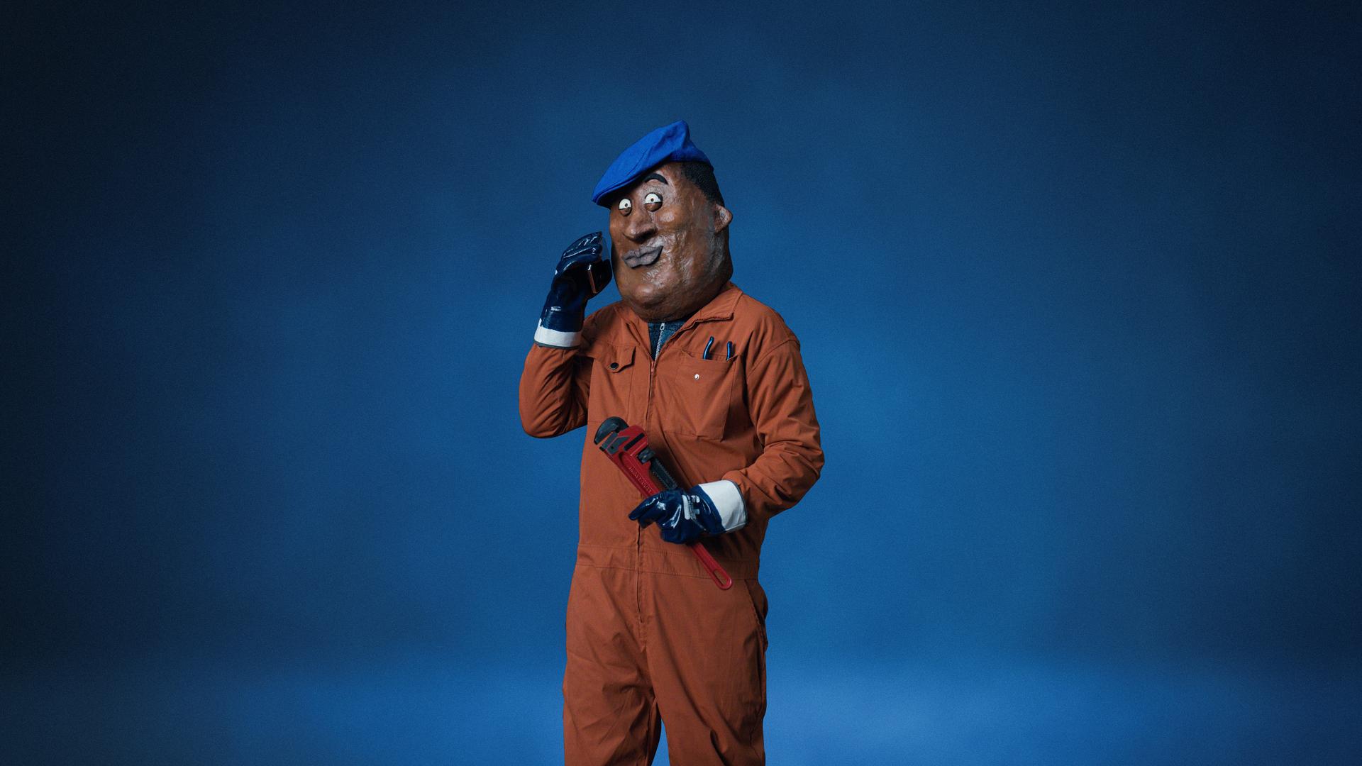 Beeld uit de video 'Bob': Medewerker in oranje werkpak en blauwe baret tegen een blauwe achtergrond.