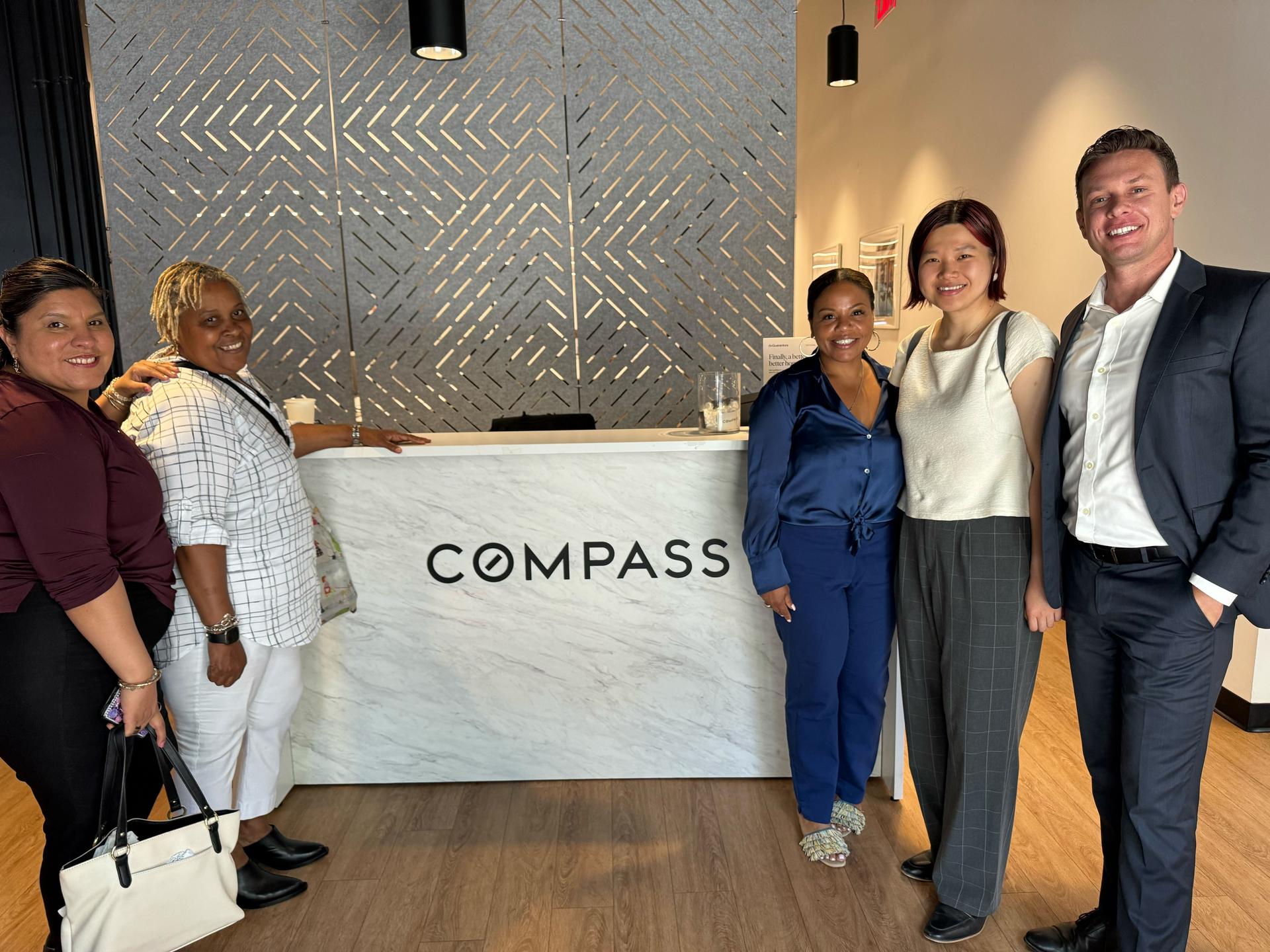 Five P E U staffers stand in front of a Compass Realty desk