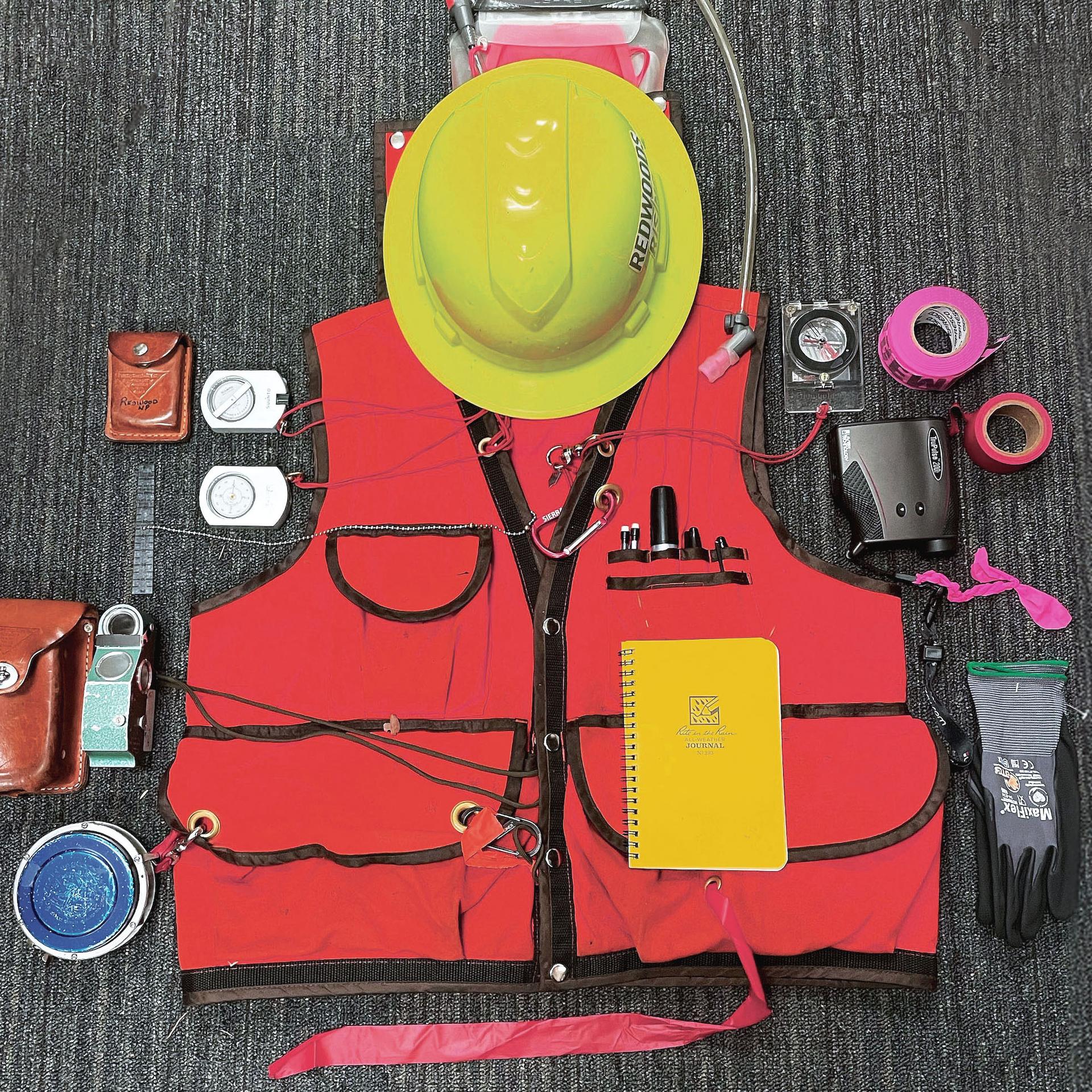Forestry gear laid out on the floor, including a vest, hard hat, compasses, and flagging tape
