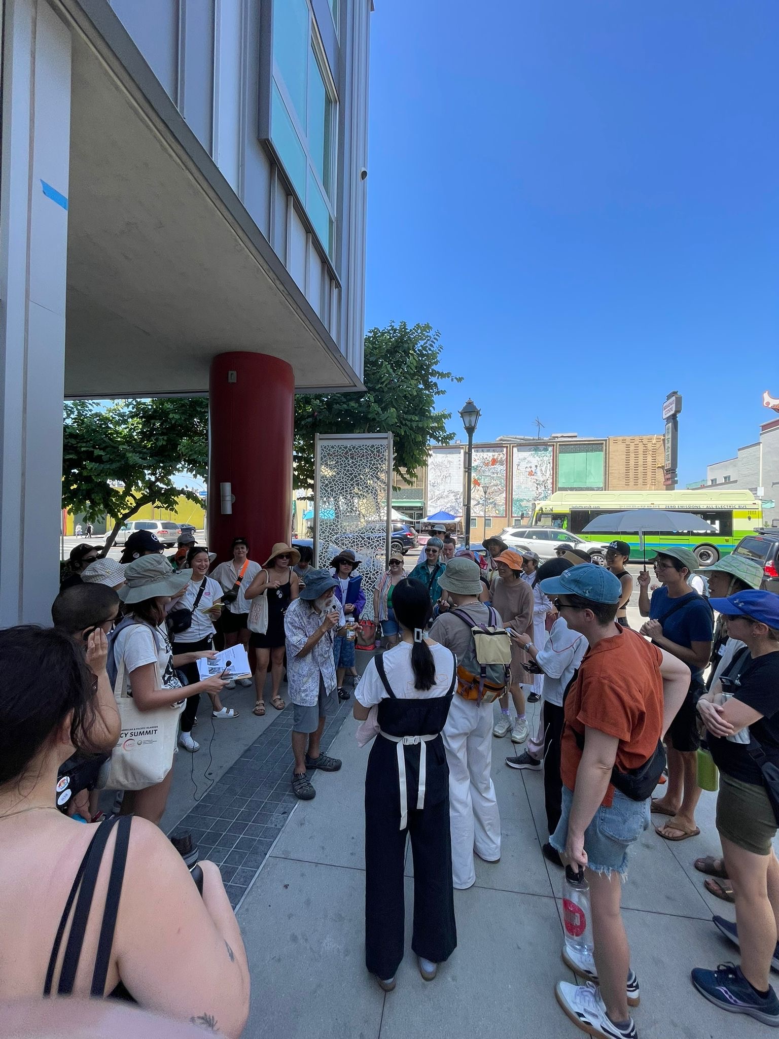 LA Chinatown community elder Eugene Moy shares knowledge about the forces shaping Chinatown into the neighborhood it is today to a group of people standing outside a large housing development.  