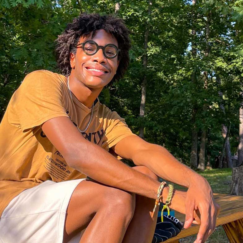 Young Black man wearing glasses sits on an outside picnic table and smiles at the camera