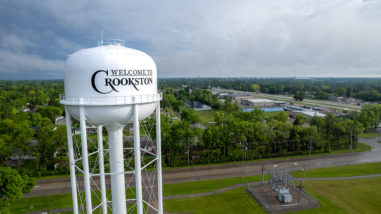 White watertower that says ''Welcome to Crookston'' with green trees, neighborhood behind