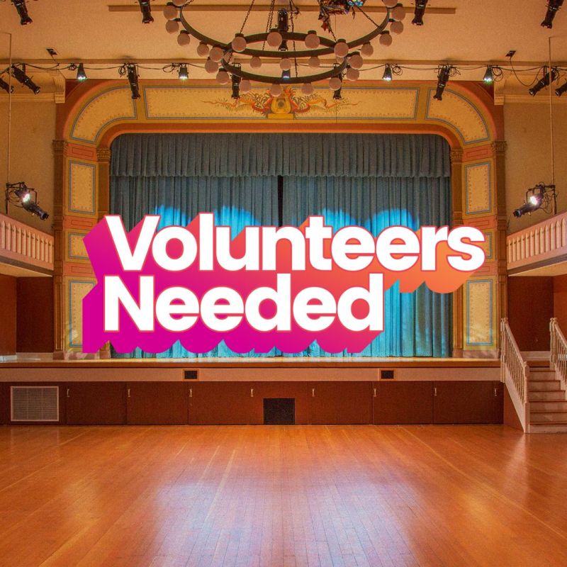  An elevated stage with wood floors and blue curtains. It reads: ''Volunteers Needed'' in white font with red shadow effect