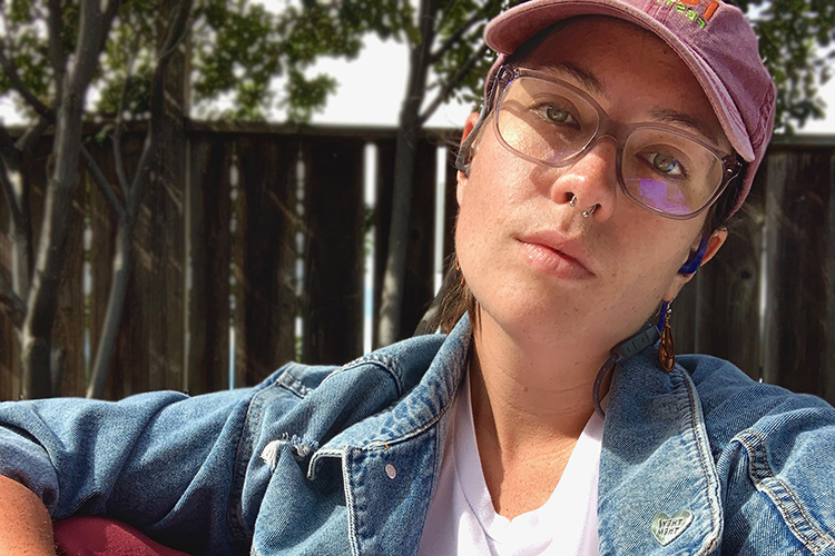 Julia facing forward wearing glasses and a pink hat outside on a sunny day with a wooden fence behind them.