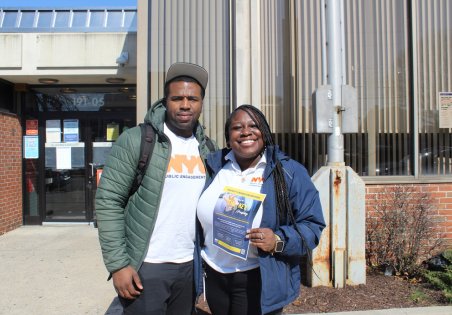 2 PEU staffers pose on the sidewalk, one holds up a flyer about housing