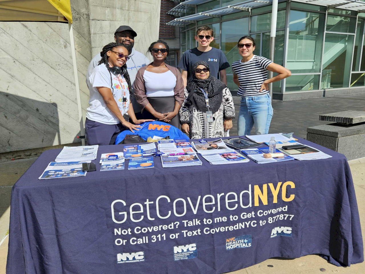 6 P E U staffers stand together at a Get Covered N Y C table with flyers piled on it. The tablecloth reads: ''Get Covered N Y C: Not covered? Talk to me to Get Covered or call 311''