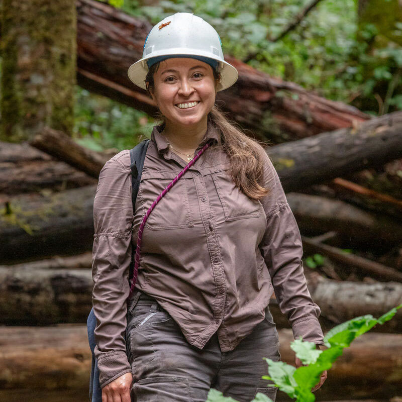 A smiling woman wearing a hard hat stands in a forest