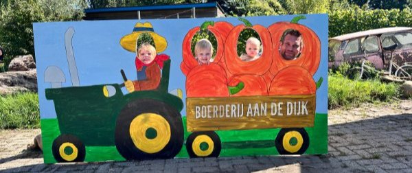 Een man en drie kinderen poseren achter de kijkgaten van een bord met daarop een trekker en kar vol pompoenen