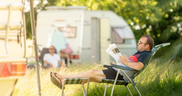 Een man zit in een tuinstoel en leest de krant met op de achtergrond een caravan