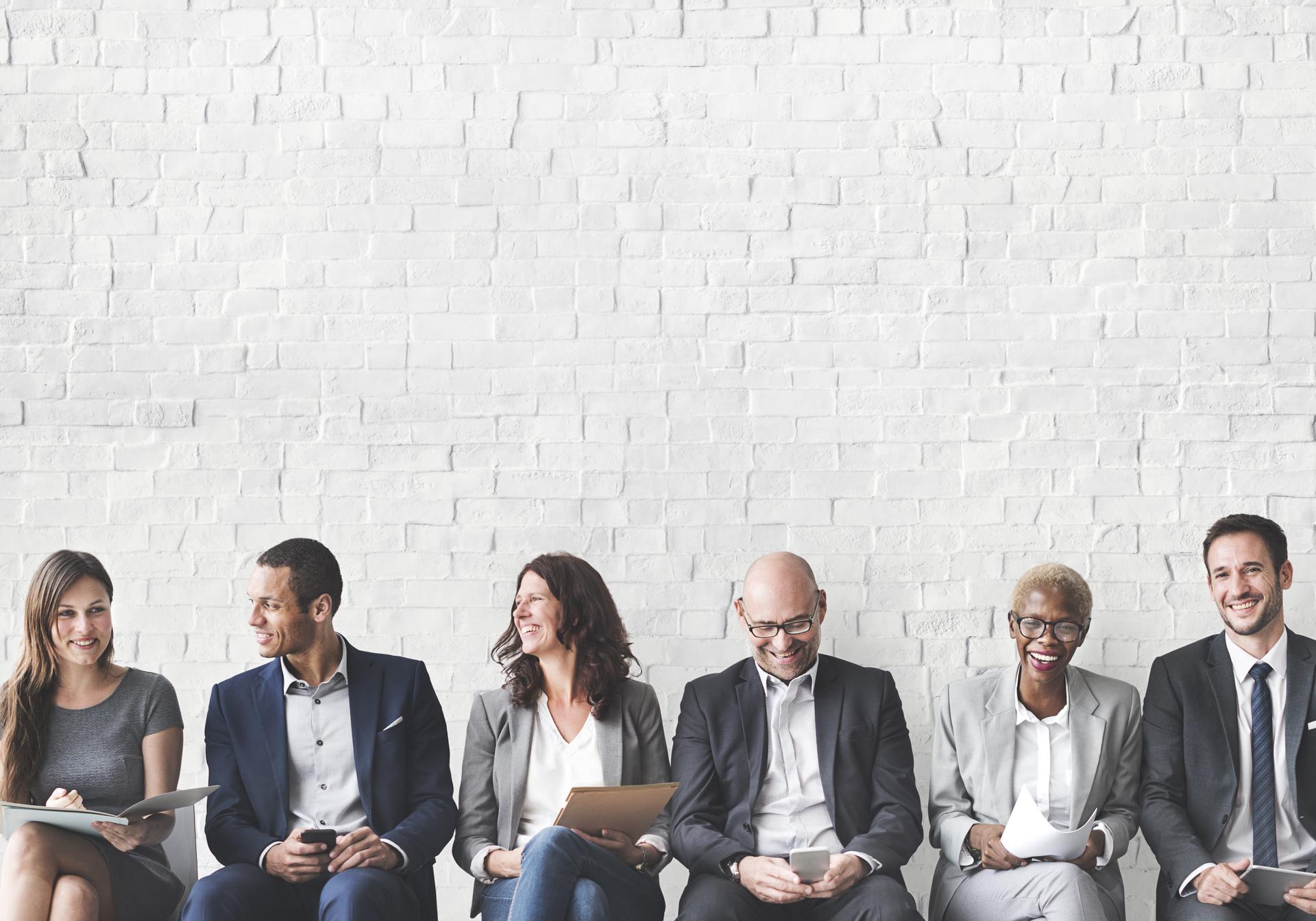 A number of professionals sitting in a row of chairs against a white brick wall