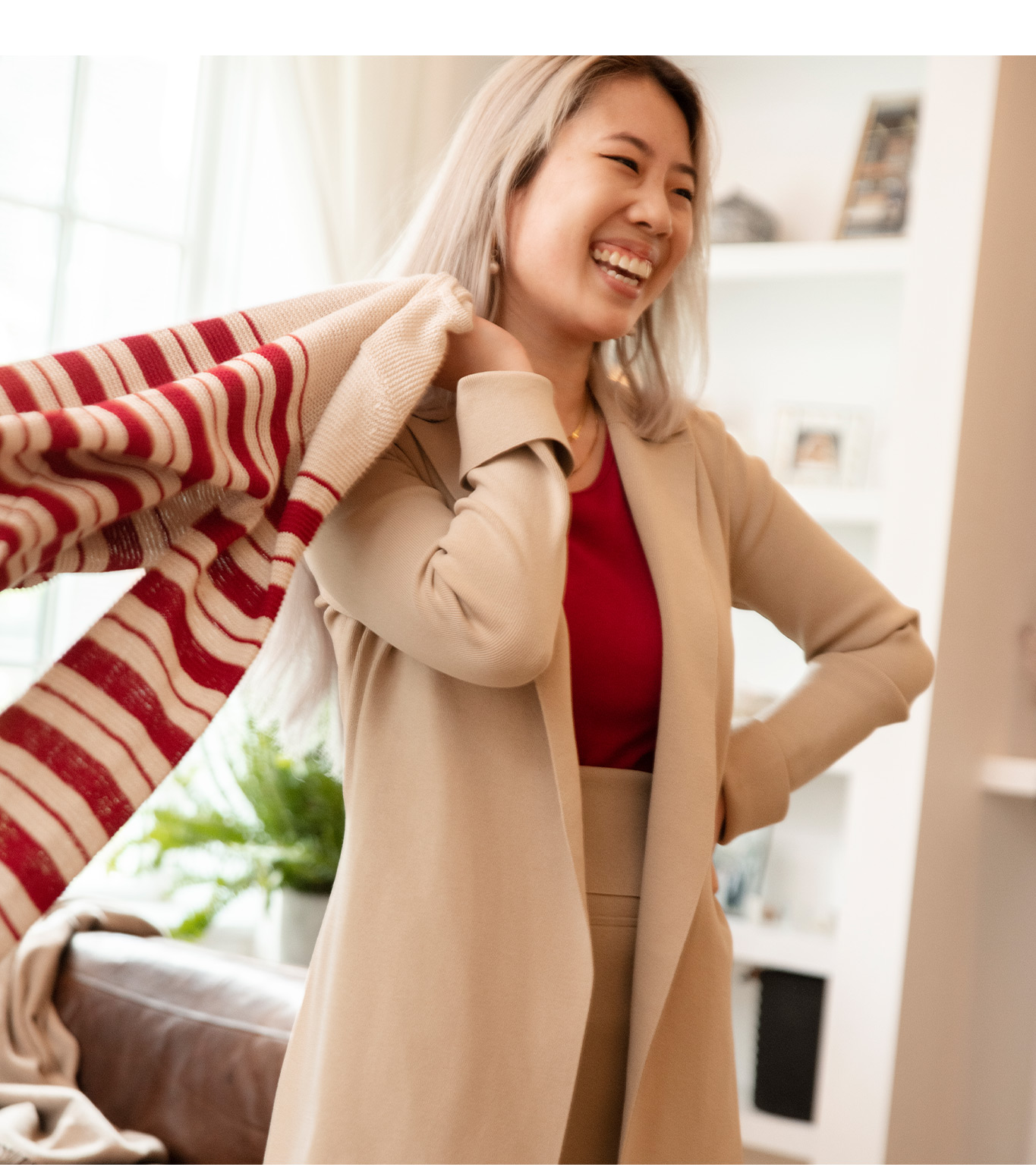 Image of a smiling woman wearing a long, beige-colored sweater over a red top and beige pants, while tossing a red-and-white-striped sweater over her shoulder.