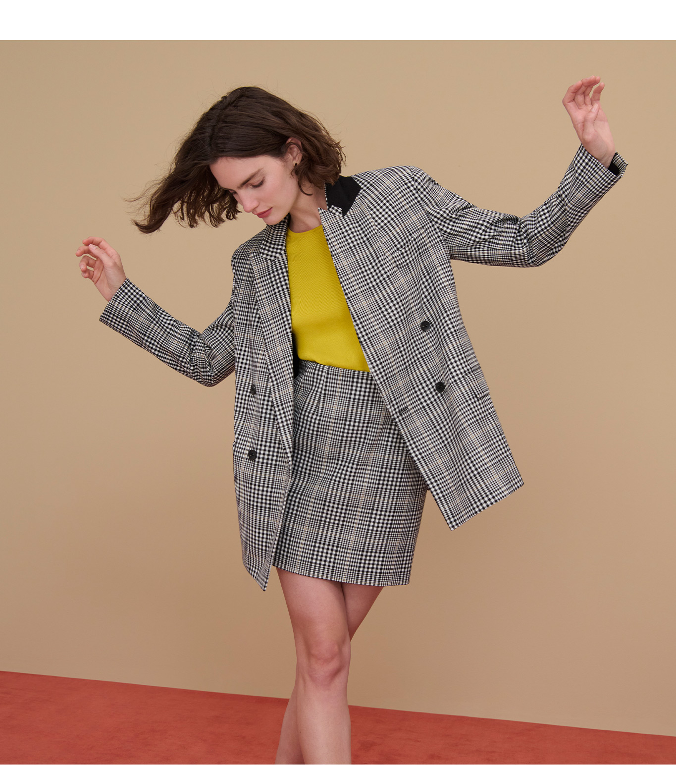 Image of a woman with short brown hair dancing on a red carpet in front of a tan wall. She's wearing a matching suit skirt and jacket in black and white plaid, and a yellow underpinning.
