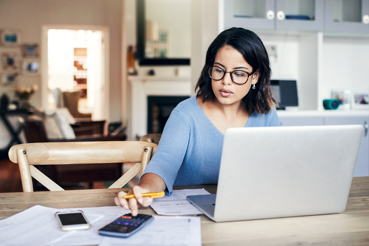 Women at her kitchen table with a computer using the calculator on her cellphone