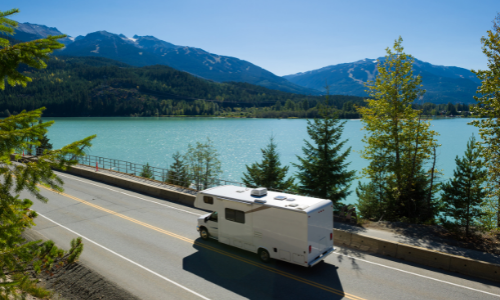 RV driving down the road in front of lake and mountains
