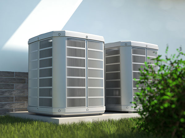 Two off-white air source heat pumps sit on a concrete pad in front of a short brick wall and behind a small bush.