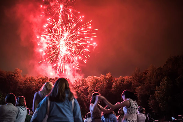 Young people dance and watch a red firework explode over a thick tree line.