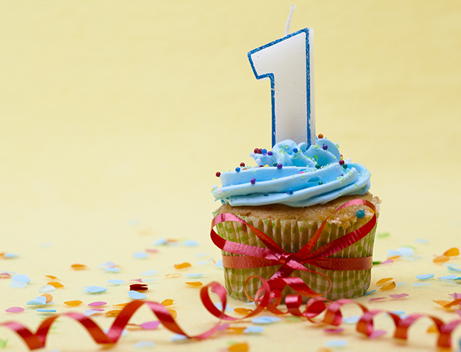 Cupcake wrapped in curly ribbon with blue frosting, sprinkles, and a 1 candle